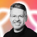 Smiling man with glasses, beard, and short hair against a vibrant orange to pink gradient background featuring a blurred white, abstract shape.