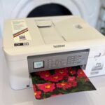 A white printer on a table printing a vibrant photo of red flowers with green leaves. The control panel and paper tray are visible.