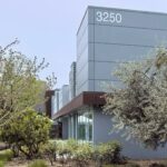 Modern gray building with the number 3250 and large windows, surrounded by trees and plants on a sunny day.