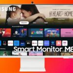 Samsung Smart Monitor M8 displaying streaming apps and family photo; white remote beside monitor on gradient orange-red background.