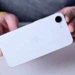 Person holding a white smartphone showing its back, featuring a camera lens and an Apple logo on a smooth, sleek surface.