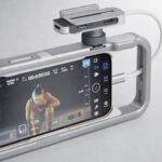 Smartphone in a gray stabilizing rig records a person kneeling, with video interface on screen and attached external device via cable.