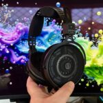 Hand holding large black Audio-Technica over-ear headphones in front of a computer monitor displaying vibrant swirling splashes of pink, purple, blue, green, and yellow paint-like colors