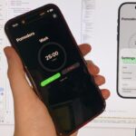 Hand holding a smartphone showing a dark Pomodoro timer app, with a large 25-minute countdown, in front of a computer screen displaying the app