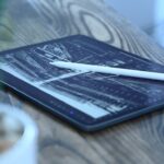 Tablet on a wooden table displaying a monochrome architectural drawing, with a white stylus resting across the screen and a blurred potted plant in the foreground