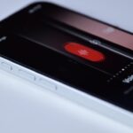 Close-up of a smartphone on a white surface displaying a recording app with a large red record button and text reading Voice Memo