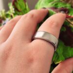 Hand wearing a wide silver ring on the middle finger, held against large green and burgundy leaves, with a thumb featuring a small abstract black-and-white nail design
