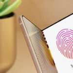 Foldable smartphone partially open displaying colorful fingerprint Touch ID screen, with a blurred potted succulent plant in the background on a warm beige surface