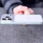 Hand holding a white smartphone horizontally above a gray surface, showcasing the side buttons and raised triple rear camera module on the back