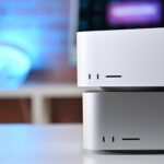 Two stacked silver Mac Studio desktop computers on a white desk, showing front vents and ports, with a blurred colorful monitor and blue light in the background