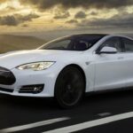 White Tesla sedan driving on an open highway at sunset, motion blurred wheels, rolling green hills and dramatic cloudy sky in the background