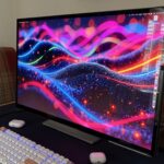 Large desktop monitor showing colorful abstract waves on a desk, with a mechanical keyboard, wireless earbuds case, and plaid-upholstered chairs in the background