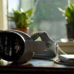 Sleek virtual reality headset and connected battery pack resting on a wooden table by a sunny window, with two small green potted plants blurred in the background