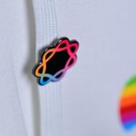 Close-up of a white fabric hoodie with a colorful gradient atom-shaped pin on the drawstring and a blurred rainbow-striped Apple logo in the background
