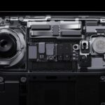 Close-up transparent smartphone back showing internal components, including a large camera lens, flash module, circuit boards, screws, and metallic parts arranged symmetrically against a dark background