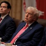 Four suited men sit at a conference table; the central man with blond hair and red tie has eyes closed, appearing contemplative, while others look forward in a formal meeting setting