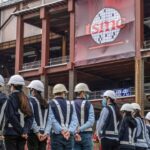 Construction workers wearing helmets and safety vests stand in a line facing a partially built factory with large TSMC banners and scaffolding in the background