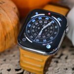 Smartwatch with orange leather strap displays analog time, surrounded by a large orange pumpkin and a smaller white pumpkin on a textured surface.
