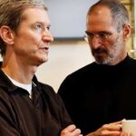 Two men are talking indoors. One holds a white mug, wearing glasses and a black shirt. The other crosses his arms, wearing a dark polo.