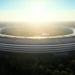 Circular futuristic office building resembling a ring, surrounded by trees at sunrise, with sunlight glowing through the center and reflecting off the smooth, layered exterior structure