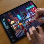 Hands using a stylus to digitally paint a colorful futuristic city street with neon signs and traffic barriers on a tablet resting on a wooden desk