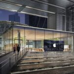 Modern glass-front Apple Store at night, surrounded by tall buildings, with illuminated cascading water steps and walkways leading to the entrance, people walking and taking photos.
