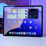 Tablet on a desk displaying colorful home screen with widgets, weather, and app icons; blurred background shows shelves and a glowing blue light sphere.