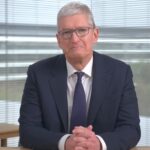 Older man with short gray hair, glasses, and navy suit sits at a desk, hands clasped, in a bright office with window blinds and potted plants in the background