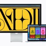 Large desktop monitor and open MacBook displaying colorful digital artwork in design software, showing bold yellow typography on the monitor and multicolored graphic stamps on the laptop screen