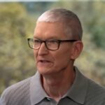 Older man with short gray hair and black glasses speaking, wearing a gray collared shirt, with a softly blurred background of green and yellow foliage behind him