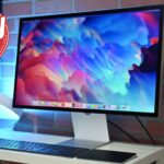 Modern Apple Studio desktop setup with large colorful monitor, slim keyboard, trackpad, headphones stand, and glowing lamp on white desk against brick wall, with red circular Daily Deal graphic in corner