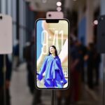 Smartphones on stands in a bright showroom, with a central phone displaying a large lock screen photo of a smiling woman in a blue outfit, background visitors blurred