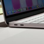 Side view of an open slim laptop showing white keyboard, two USB-C ports on the left edge, and colorful app icons on the lower part of the display