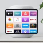 Smart speaker with a tablet-like screen displaying colorful streaming and media app icons, including Apple TV, Netflix, YouTube, and CBS, on a white table beside a green houseplant