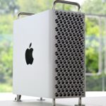 Silver Apple desktop tower with perforated front panel, chrome handles and feet, standing on a white desk near a large window overlooking a green, blurry outdoor background.