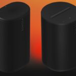 Two tall black Sonos wireless speakers with rounded edges and touch controls, standing side by side against a warm orange to black gradient background
