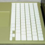 Open slim laptop with light green body, large trackpad, and white keyboard, viewed from above at an angle, screen dark and reflecting the keys on a gray surface