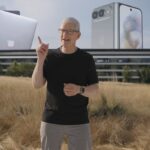 Man in black shirt presenting outdoors before Apple Park, with large floating graphics of MacBooks and iPhones behind him under a clear sky