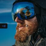 Bearded skier wearing helmet and reflective goggles against bright blue sky, seen close up, with a computer cursor and small editing markers scattered across his face and gear