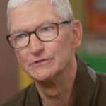 Older man with short gray hair and glasses speaking, shown in close-up indoors with a softly blurred, colorful background.