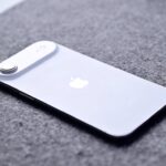 Silver iPhone lying face down on a gray fabric surface, showing the Apple logo and raised rear camera in the top left corner