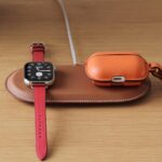 Apple Watch with red leather band and orange leather AirPods case on a brown wireless charging pad, resting on a light wooden desk with a metal keyring attached