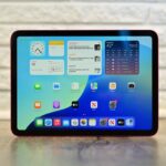 Tablet with colorful home screen showing clock, calendar, weather, and app icons, standing upright on a wooden surface against a light brick wall background