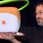 Man in black turtleneck proudly holding an orange and white Apple iBook laptop on stage, with a glowing green ring shape in the dark background