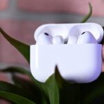 AirPods Pro 3 earbuds in an open charging case resting on green plant leaves, with a pink badge on the left showing a percentage symbol and the words PRICE DROP