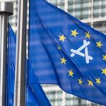 Blue European Union flag waving on tall metal pole, yellow stars in a circle surrounding stylized white airplane symbol, with modern glass office building in the background