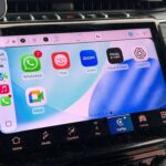 Car dashboard touchscreen running Apple CarPlay, showing WhatsApp, Yoto, Zoom, Focal & Naim, ChatGPT, and Meet apps, with climate controls and navigation buttons below in a modern vehicle interior