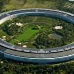 Aerial view of a huge circular office building surrounding a lush central park with trees, walking paths, a pond, and a colorful rainbow arch sculpture