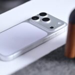 Silver smartphone lying face down on a white surface, showing a triple camera bump and smooth back, with part of a black and copper object visible on the right side