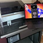 Large enclosed QIDI 3D printer on a wooden table, with a laptop displaying colorful robotic artwork and printer control software, beside a window with blinds and a nearby door